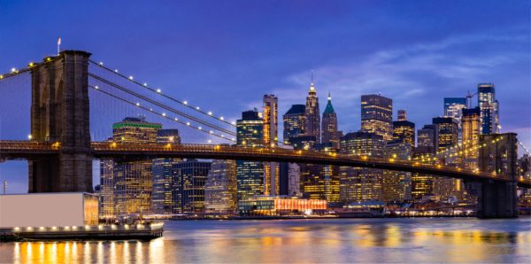 Evening view of the Brooklyn Bridge and New York City skyline with lights reflecting on the river behind it.