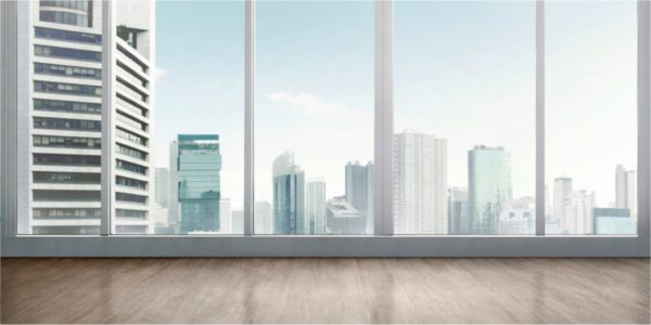 Empty room with large floor-to-ceiling windows overlooking a city skyline of tall glass buildings on a sunny day.