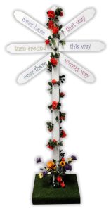 White six-arm signpost with phrases 'Over here', 'that way', 'turn around', 'this way', 'over there', 'wrong way' on a rose-covered post.